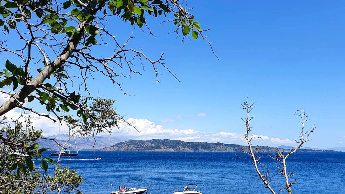 Agni Bay in Corfu, seen from the hillside through olive branches, with motorboats anchored in turquoise water and the mainland mountains beyond