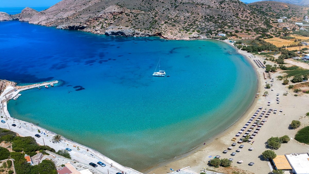 Aerial view of Galissas Bay on the west coast of Syros: a horseshoe-shaped sandy beach wrapped around turquoise shallows, a white sailboat anchored in the deeper blue water at the mouth of the bay, sunbeds along the shore with the village and parking set back behind the sand, and bare Cycladic hills rising on either side